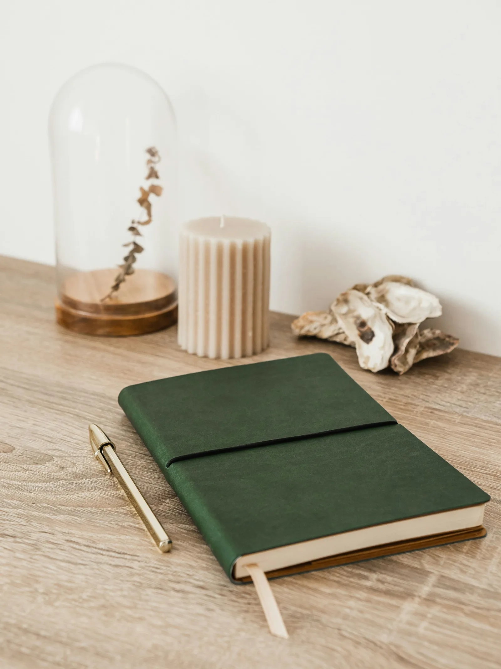 A closed green soft-cover notebook on an oak desk, a brass pen beside it.