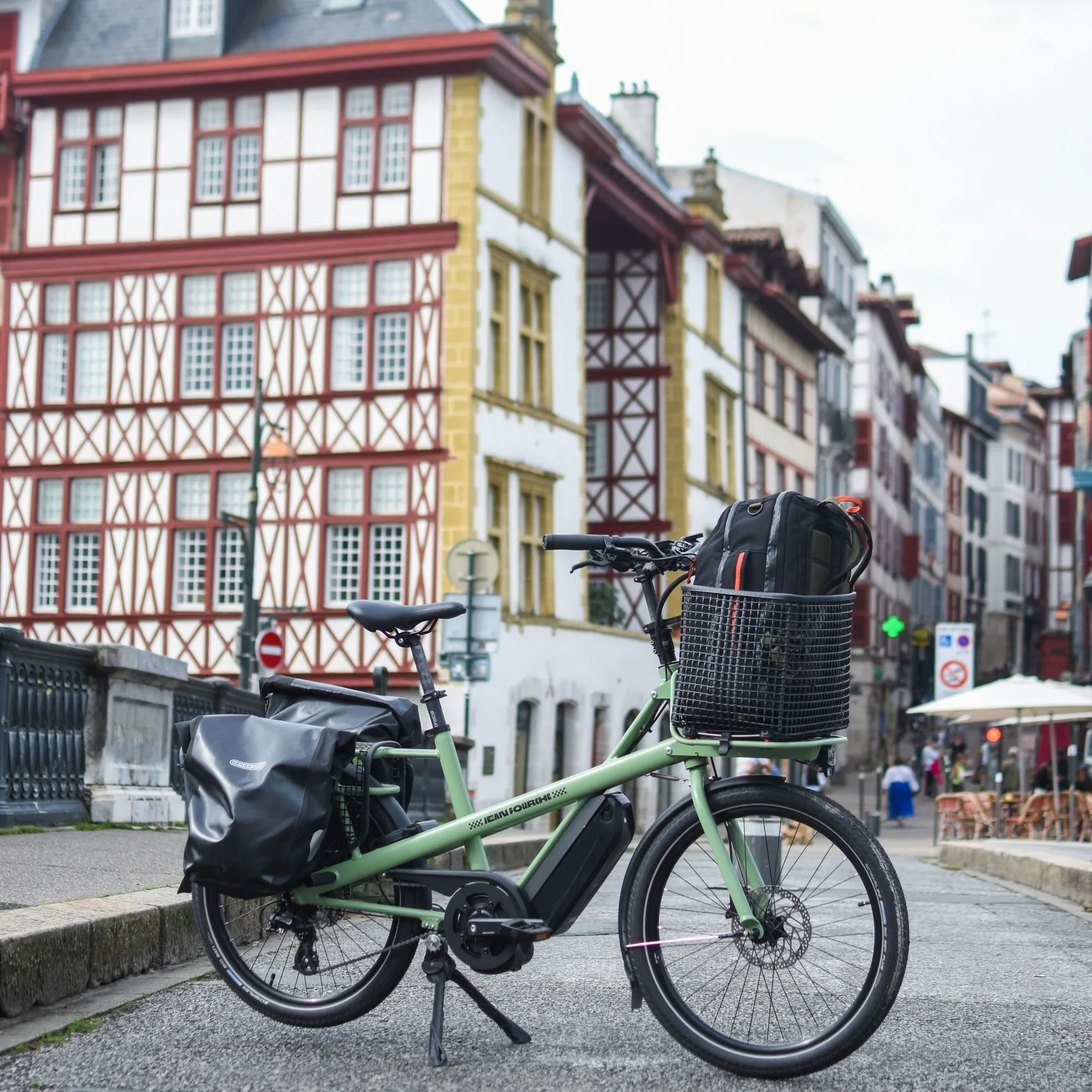 Sage green e-bike with a front basket on a cobbled street