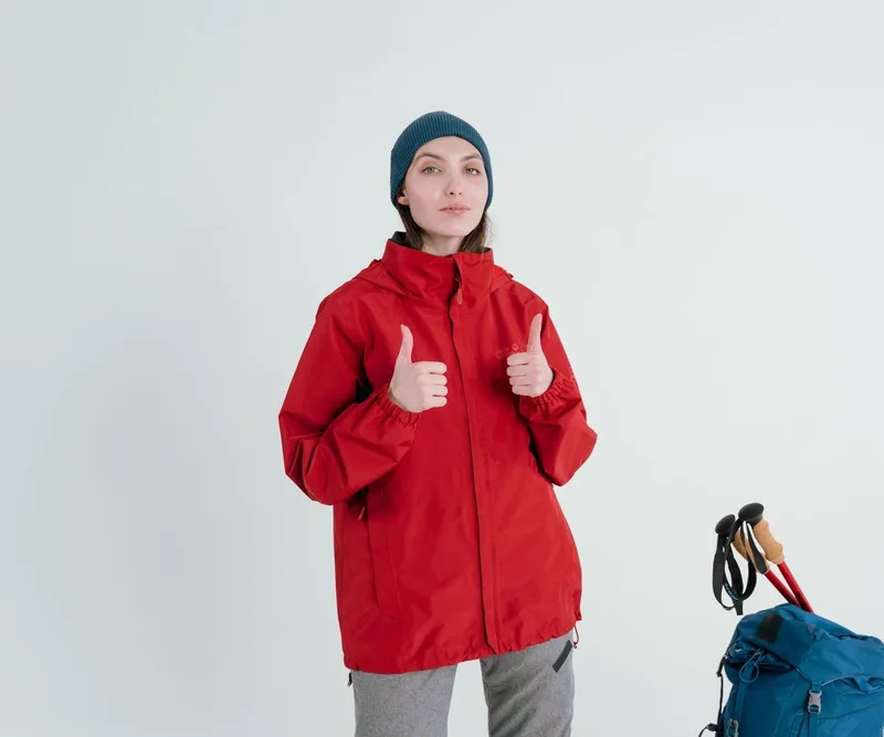 A hiker in a red waterproof jacket beside a blue backpack and poles